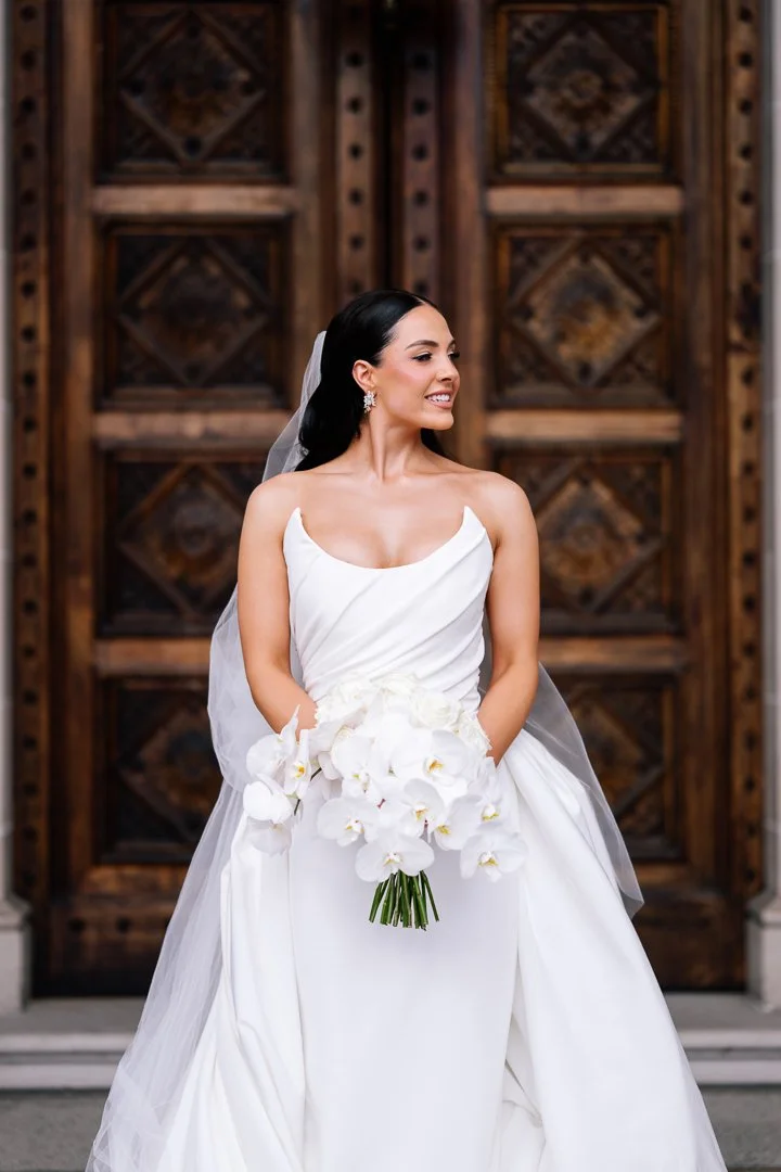 A bride in a white wedding gown holding a bouquet of white orchids, standing in front of a wooden door.