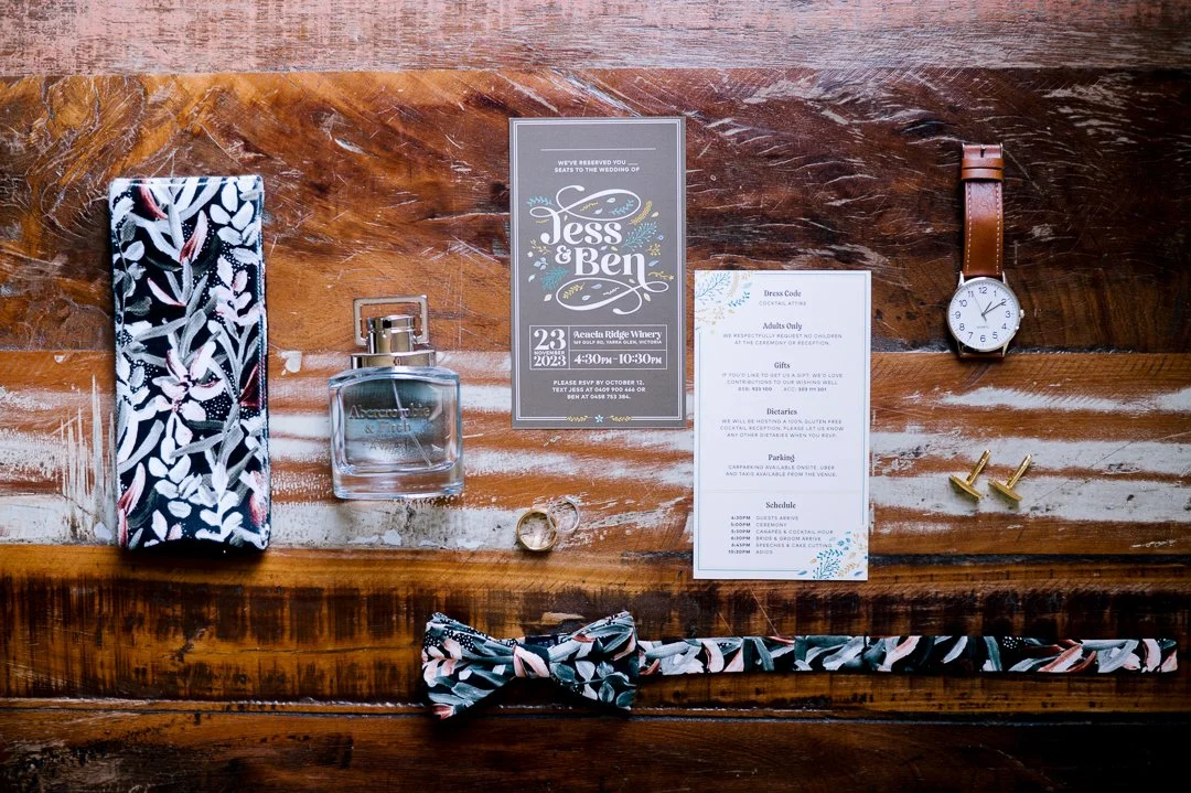 Wedding invitation with rings, watch, bow tie, perfume, cufflinks, and a fabric item on a wooden surface.