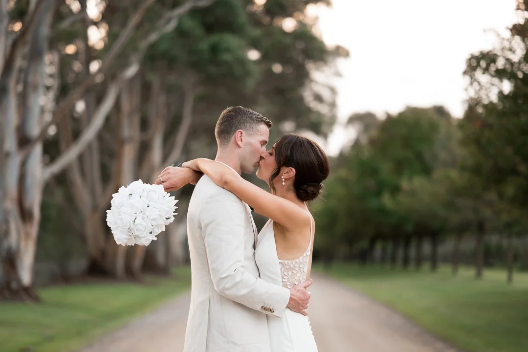 A newlywed couple sharing a kiss outdoors on a dirt road lined with trees, the woman holding a bouquet of white flowers.