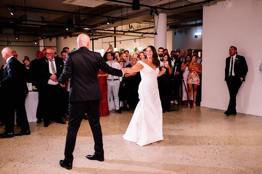 A bride and groom are dancing at their wedding reception, surrounded by guests in a modern, open hall.
