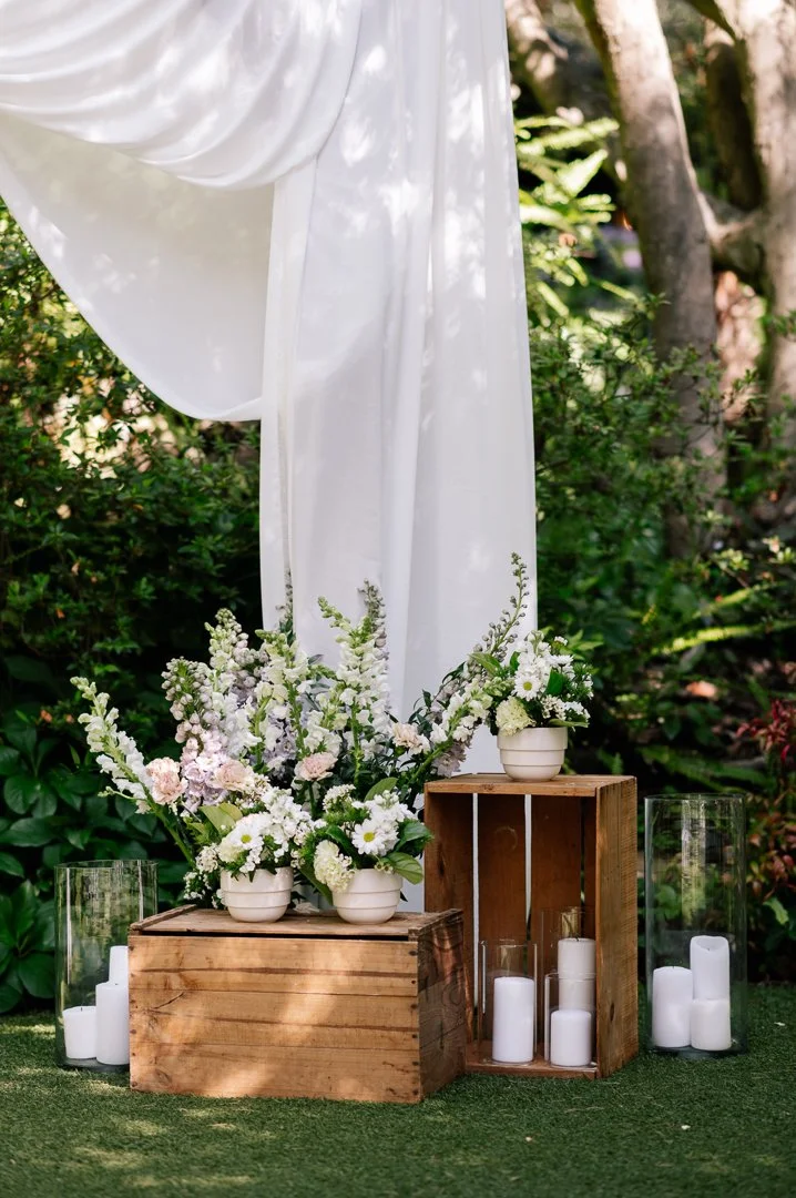 Floral decorations with white and light pink flowers in white pots, wooden crates, and candles in glass holders outdoors with greenery and a white draped fabric backdrop.