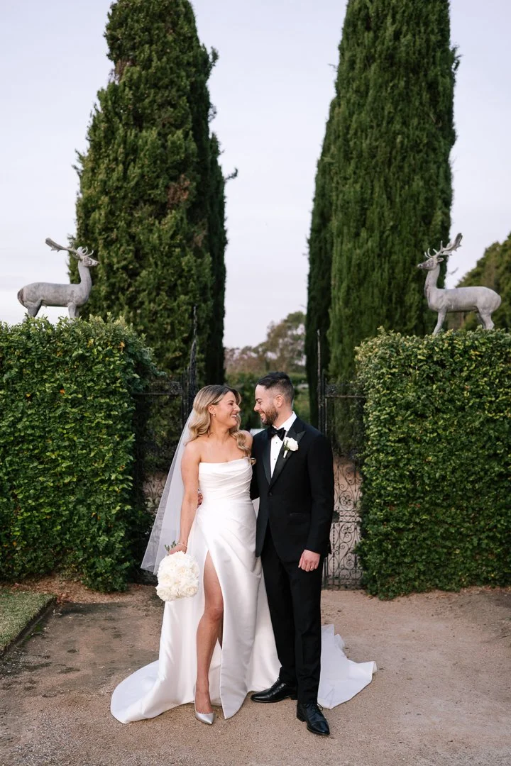 A bride and groom in wedding attire standing close together, smiling at each other in an outdoor garden setting with tall cypress trees and decorative reindeer statues in the background.