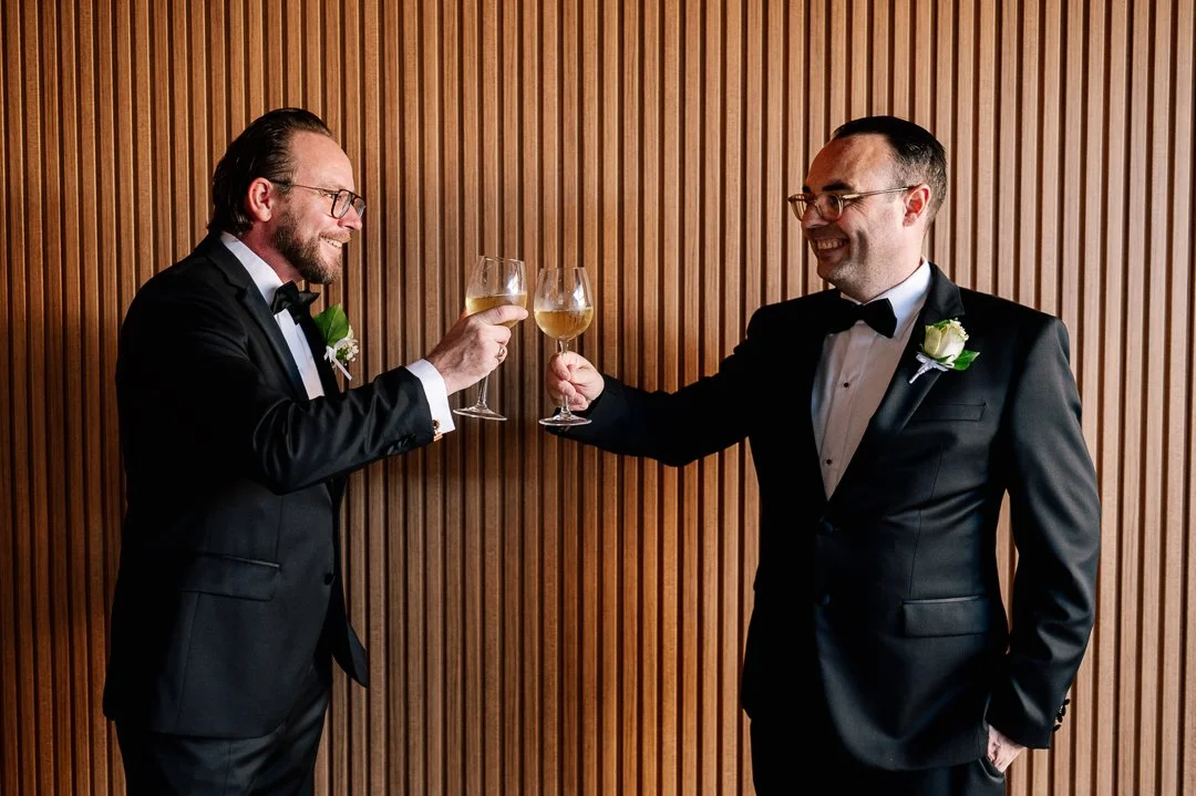 Two men in tuxedos with boutonnières clink glasses of champagne in celebration against a wooden paneled wall.