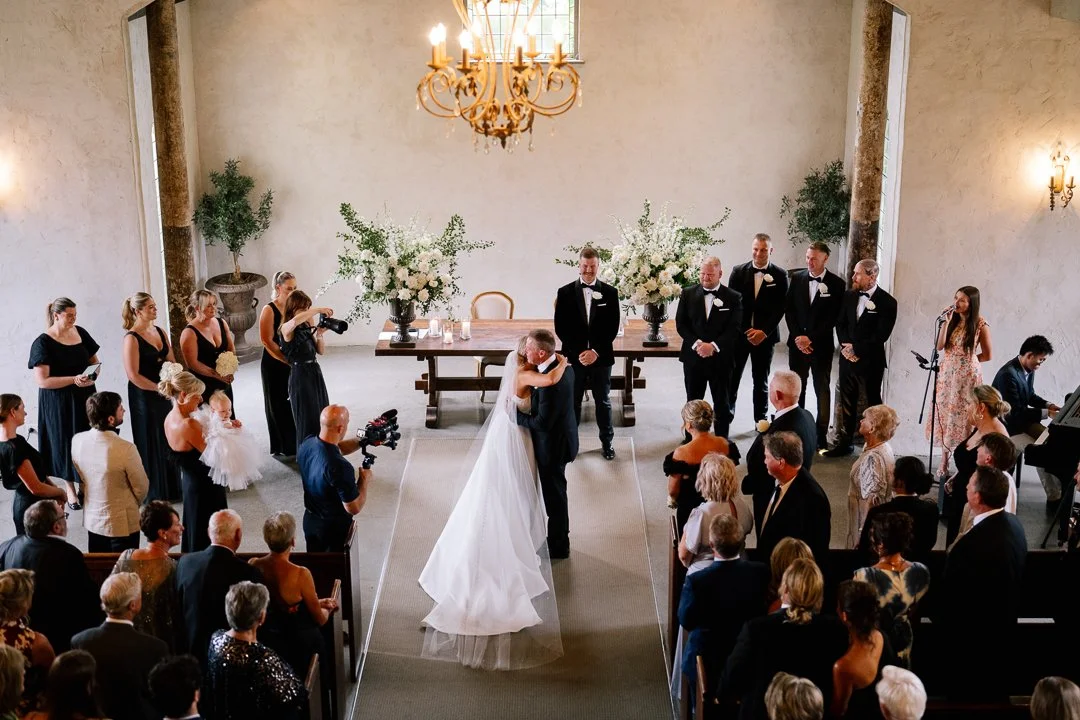 A wedding ceremony inside a church with the bride and groom kissing at the altar, surrounded by guests and bridesmaids and groomsmen standing on either side.