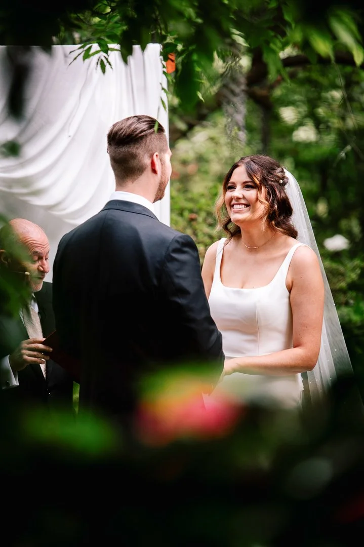 A bride and groom exchanging vows at an outdoor wedding ceremony surrounded by greenery, with an officiant speaking nearby.