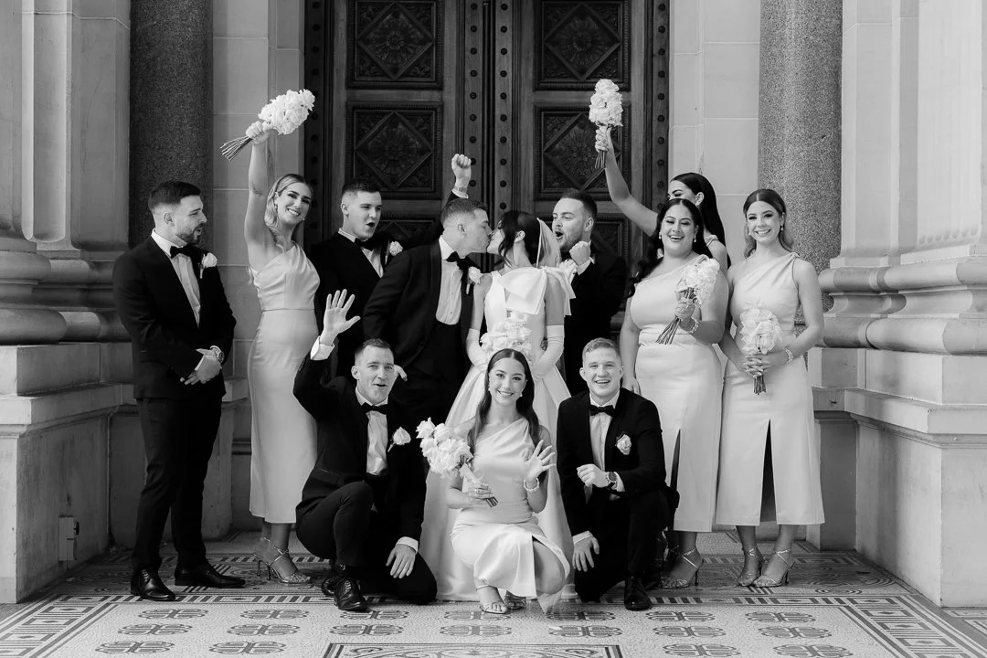 Black and white wedding photo of a group of twelve people, including the bride and groom, posing in front of an ornate wooden door and stone columns. The bride is seated with the groom kneeling beside her, all dressed in wedding attire, surrounded by