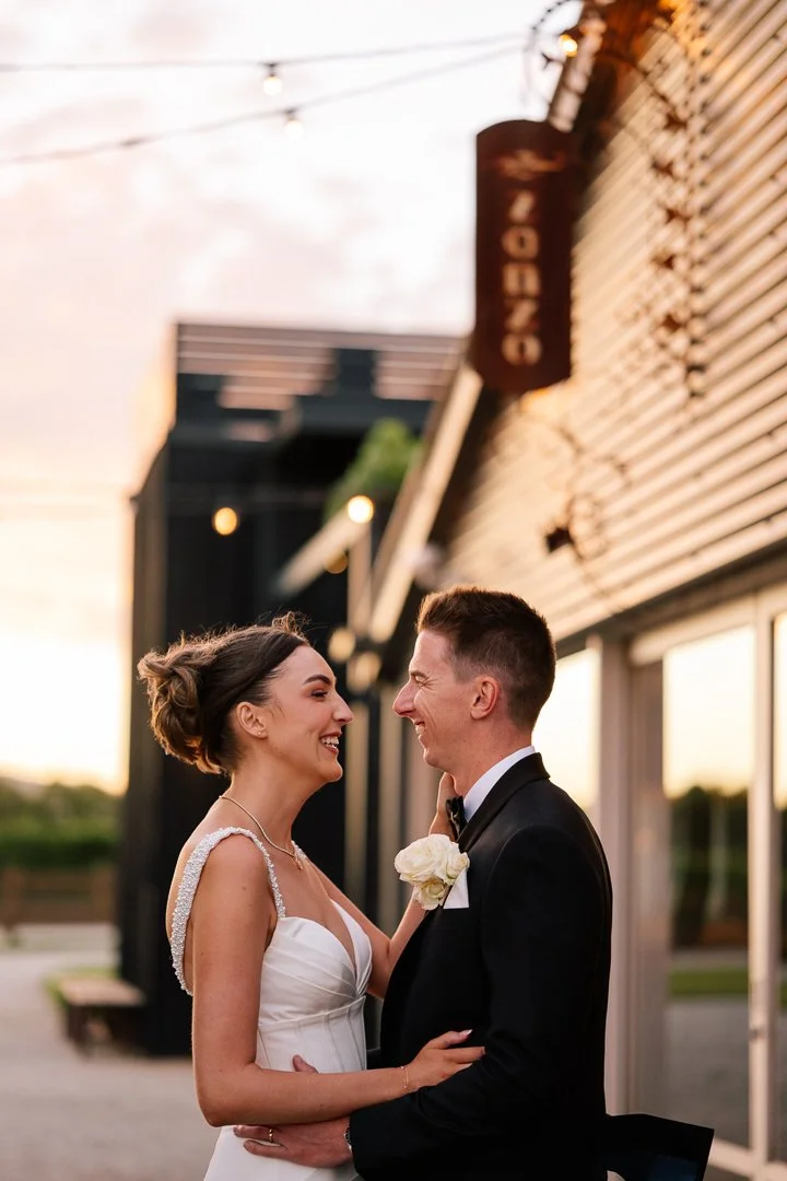 A newlywed couple smiling at each other outdoors at sunset, dressed in wedding attire, with the woman wearing a white gown and the man in a black tuxedo, standing near a building with a sign reading 'EATON' vertically.