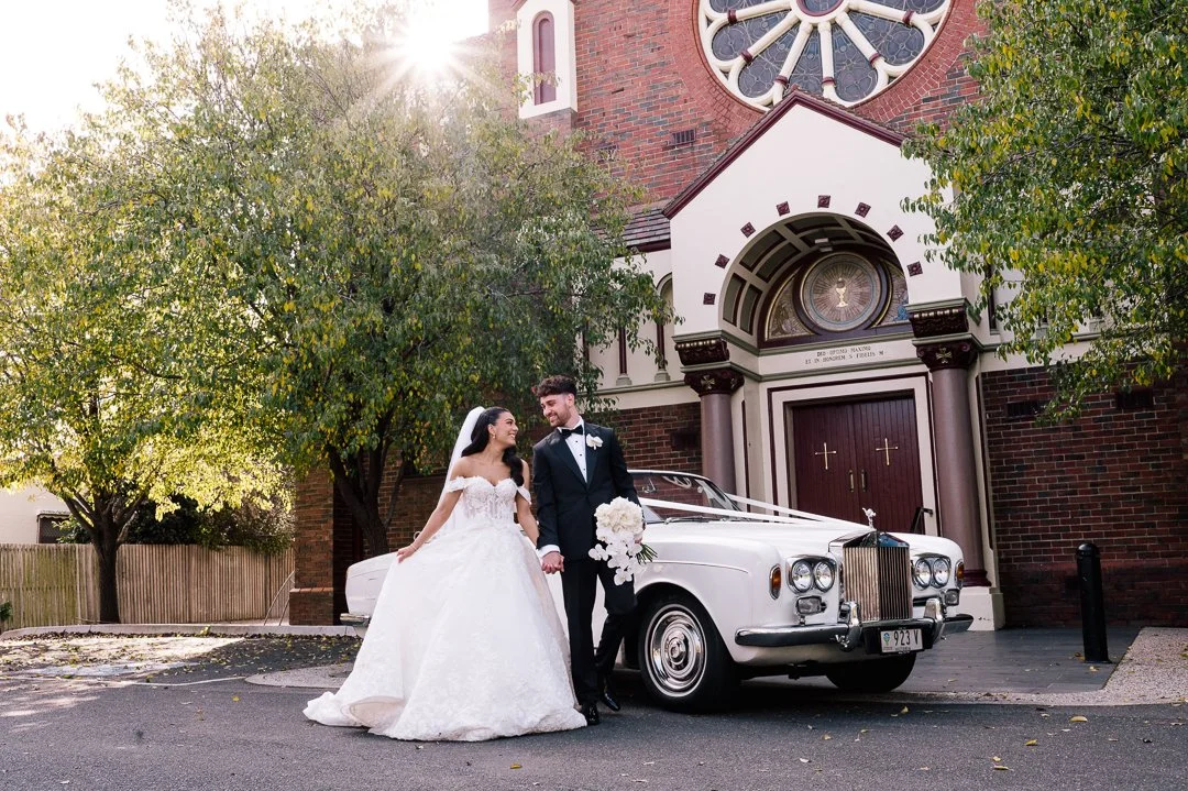 A bride and groom in wedding attire standing in front of a white luxury car decorated with a ribbon, outside a church with a large circular stained glass window, trees, and greenery.