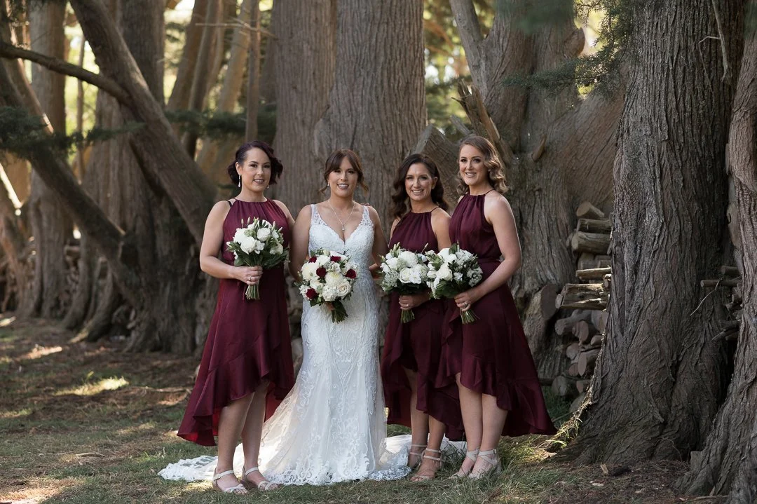 Four women standing outdoors among tall trees; one woman in a white wedding dress and three bridesmaids in burgundy dresses, each holding a bouquet of flowers.