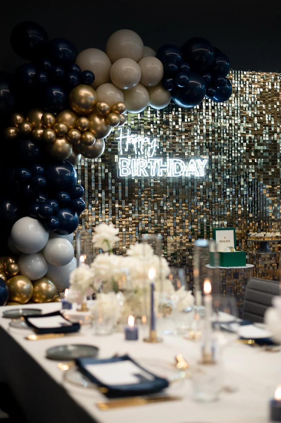 Elegant birthday celebration with a decorated table, floral centerpieces, and a balloon arch in black, white, and gold, against a mosaic wall with a neon 'happy birthday' sign.