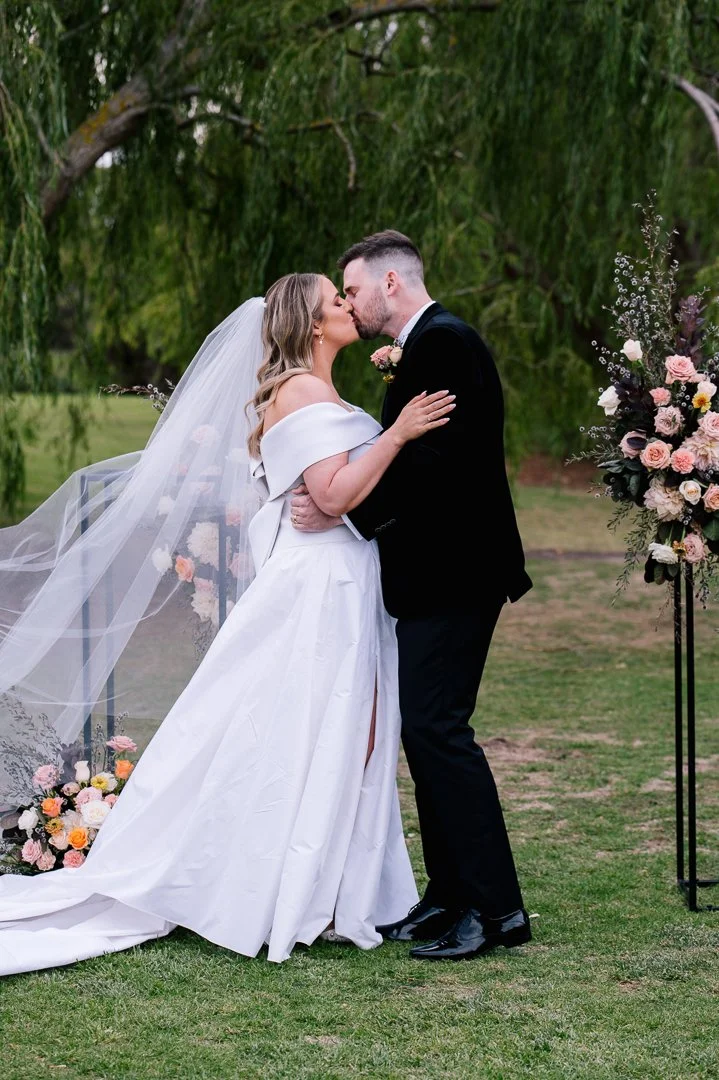 A bride and groom kiss during their outdoor wedding ceremony, decorated with floral arrangements and greenery.