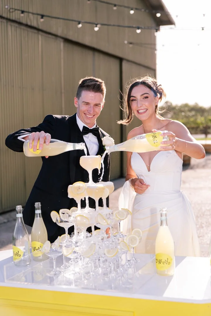 A newlywed couple in formal wedding attire pouring lemonade into a pyramid of glasses with lemon slices during their wedding celebration outdoors.