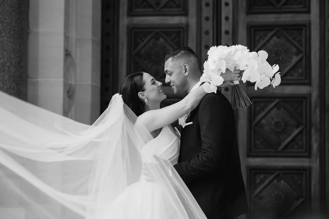 Black and white photo of a bride and groom embracing outside a building with a detailed wooden door. The bride is holding a bouquet of white flowers, and they are smiling at each other.