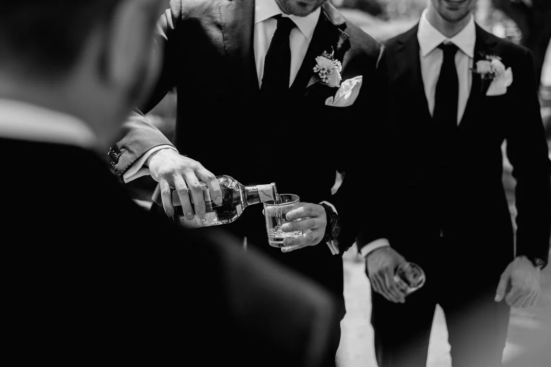 Groom pouring a drink for a guest at a wedding, all wearing suits with boutonnières.