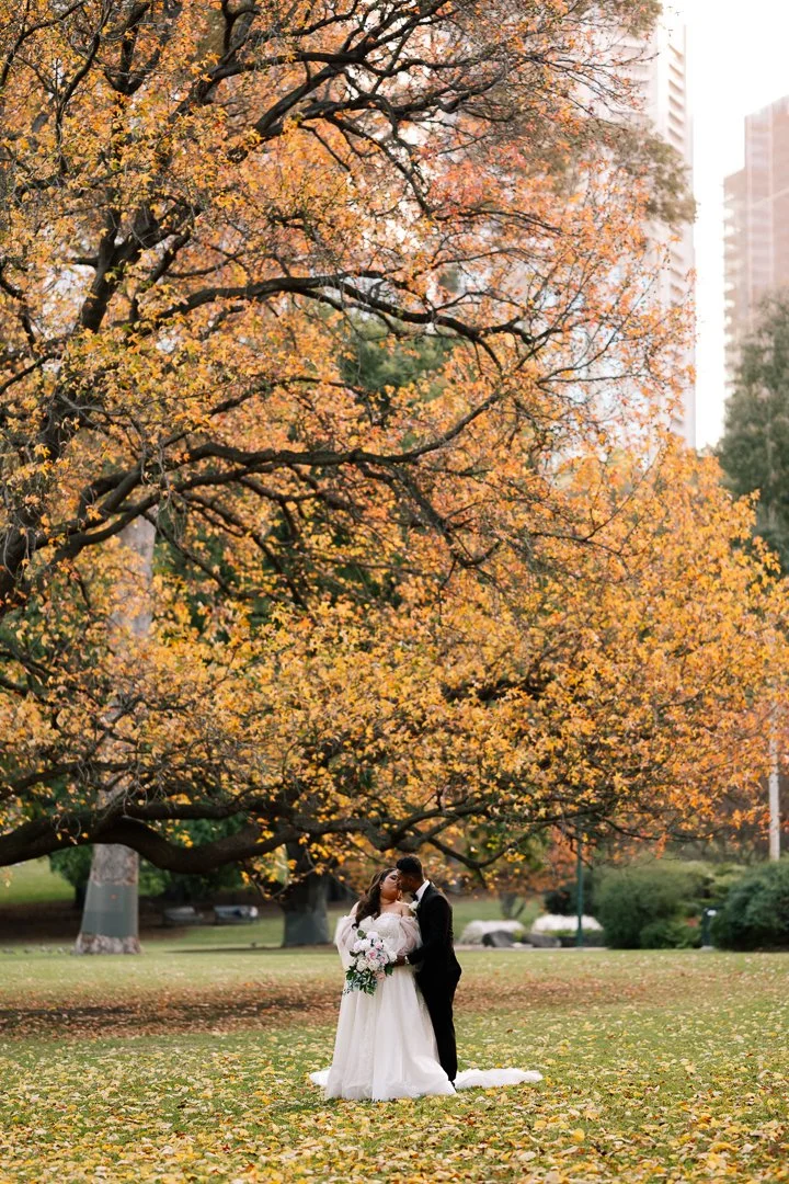 A bride and groom standing together in a park with colorful autumn leaves on a large tree above them. The bride is holding a bouquet and is wearing a white wedding gown, while the groom is in a black suit.