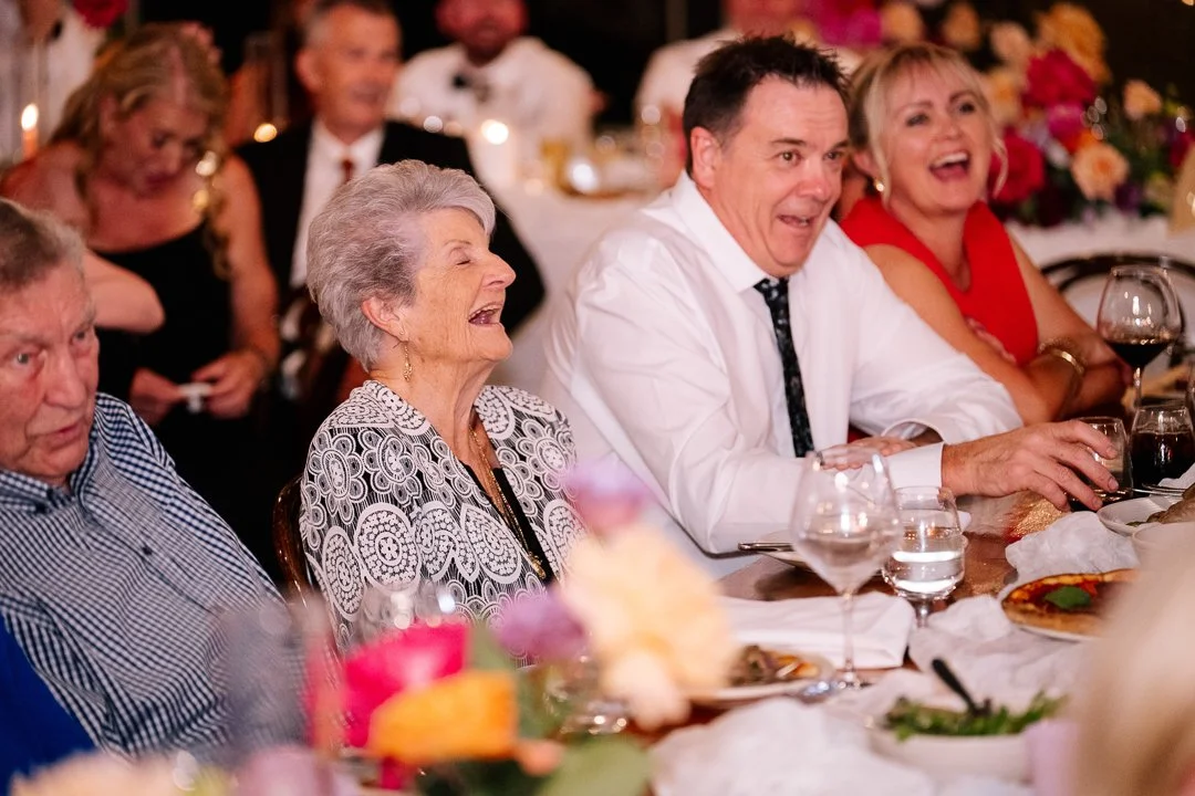 People sitting at a dining table laughing and enjoying a celebration, with food, wine glasses, and floral decorations.