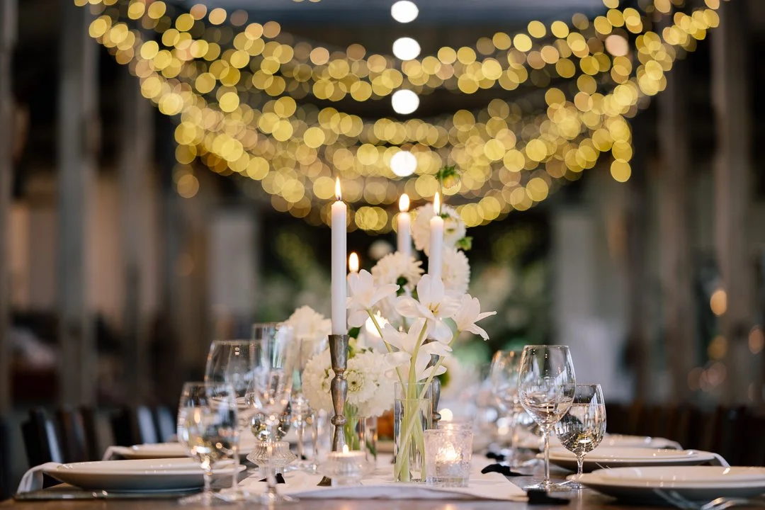 Elegant table setting with white flowers and candles under hanging string lights at night.
