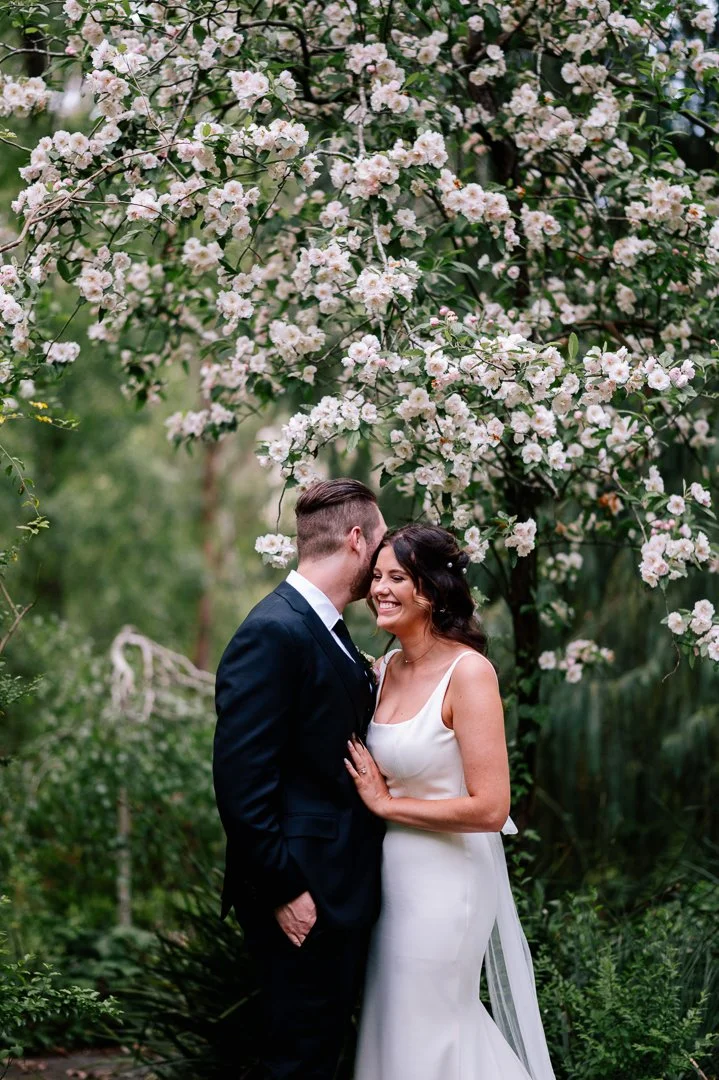 A bride and groom standing close together under a blooming tree, smiling happily at each other, in a lush outdoor setting.