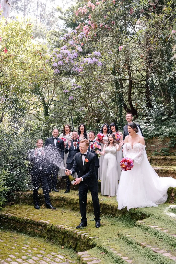A wedding celebration outdoors with a bride, groom, and bridal party on moss-covered steps surrounded by flowering trees, as the groom opens a champagne bottle with champagne spraying out.