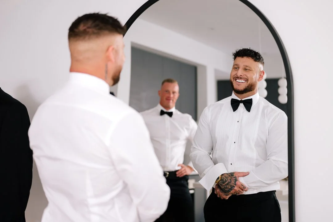 Three men in white dress shirts and black bow ties admire their reflection in a mirror, smiling and preparing for a formal event.