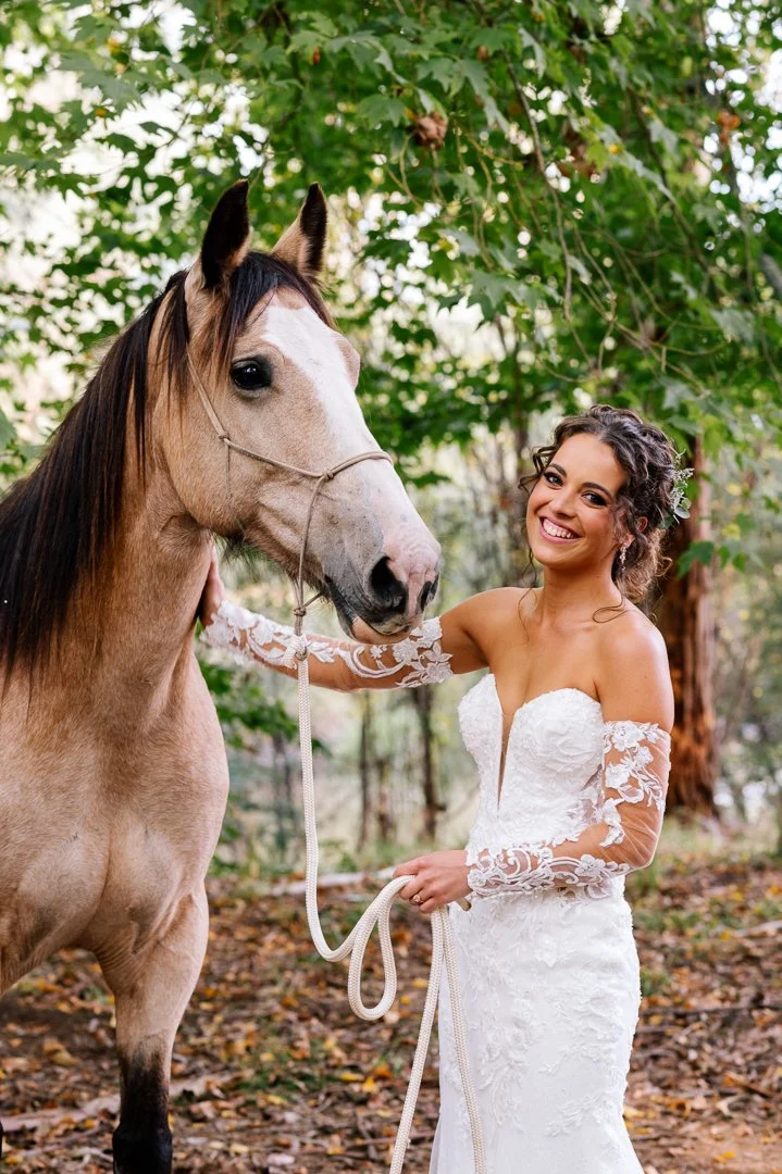 A woman in a white wedding dress with lace sleeves standing outdoors holding a rope, smiling, next to a light-colored horse with a bridle, surrounded by trees and green foliage.