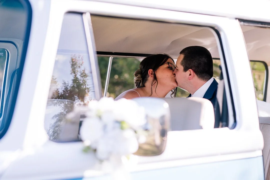 A newlywed couple sharing a kiss inside a white vehicle, with blurred flowers and greenery outside.