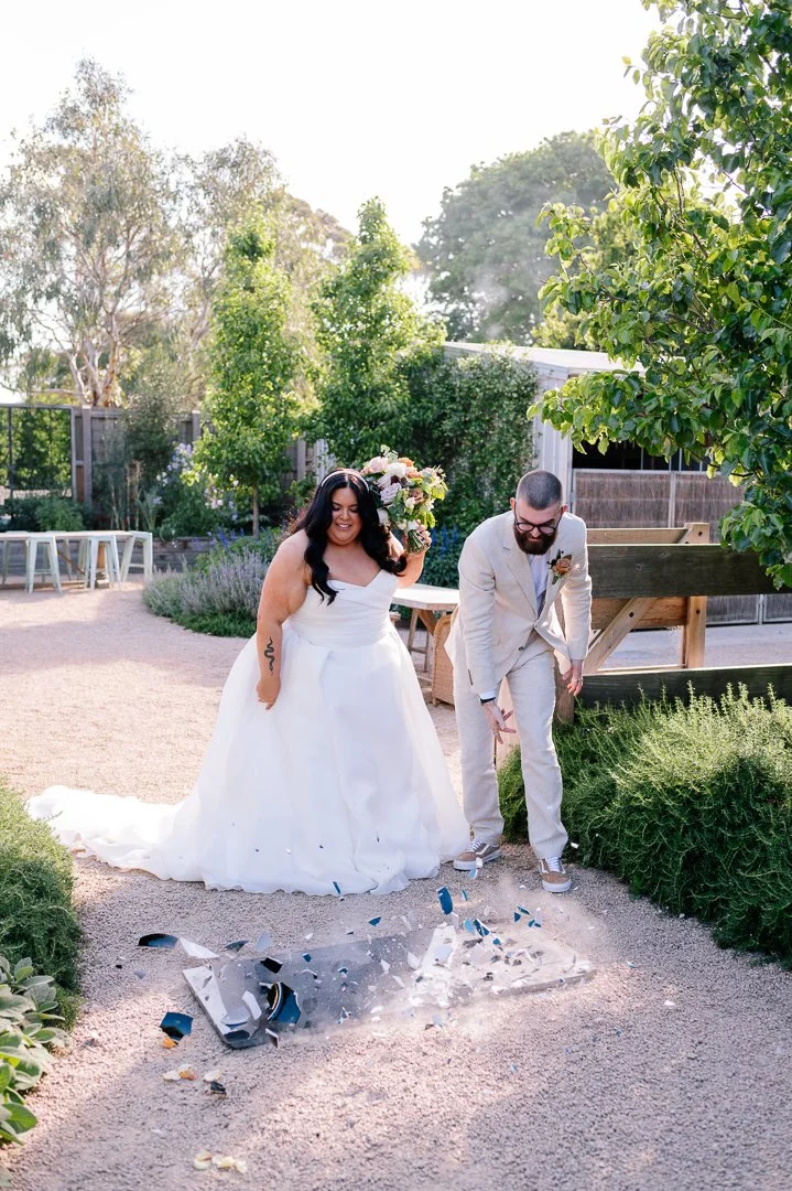 Wedding celebration outdoors with a bride in a white gown and a groom in a light-colored suit breaking a glass on the ground, surrounded by greenery and sunlight.