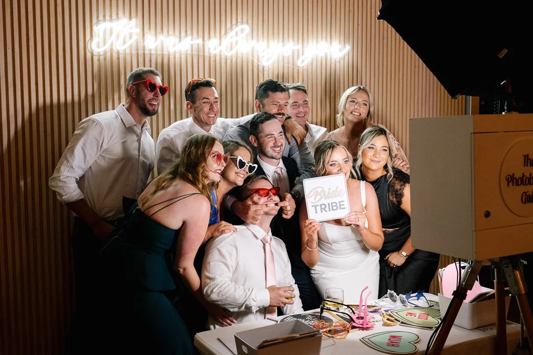 Group of people at a wedding or celebration party taking a photo, some wearing sunglasses, holding signs, with a neon sign in the background.