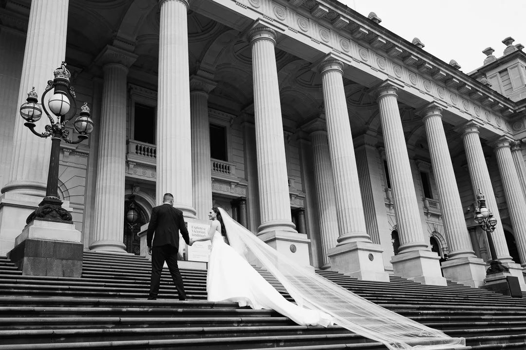 A bride and groom holding hands on grand stairs in front of a classical building with tall columns, ornate street lamps, and arched windows.