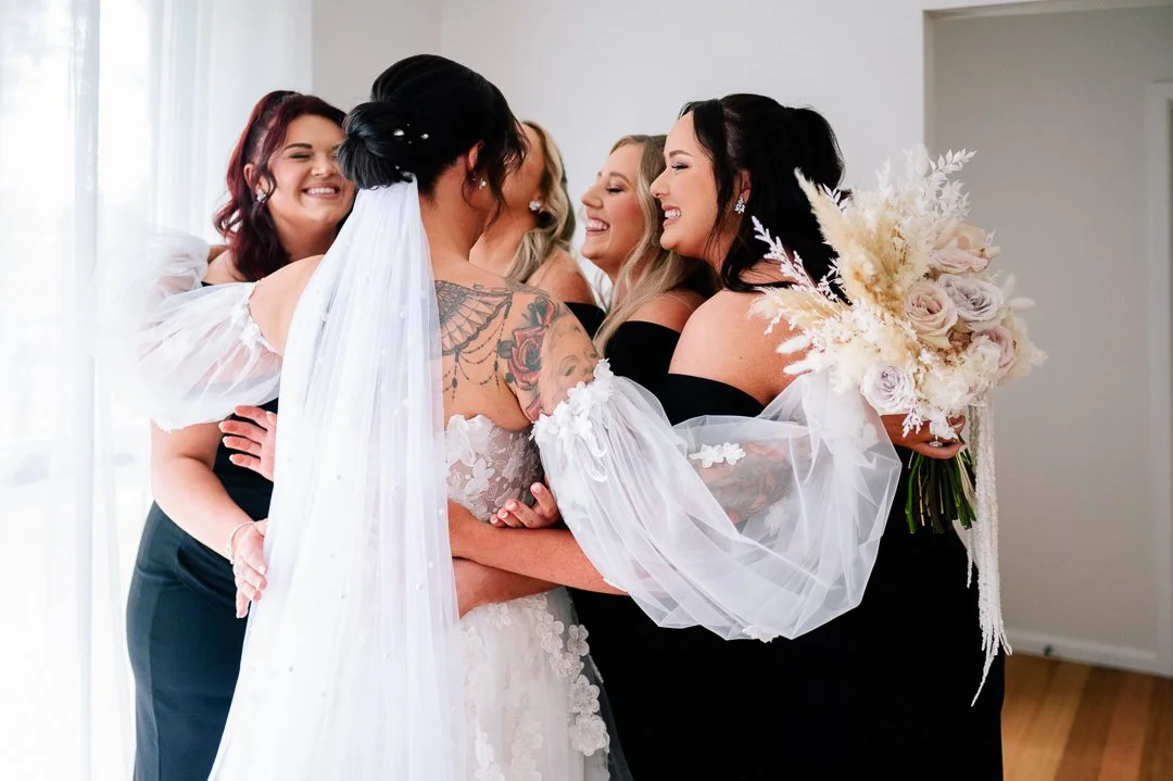 Bride and bridesmaids hugging indoors, bride holding a bouquet, all smiling.