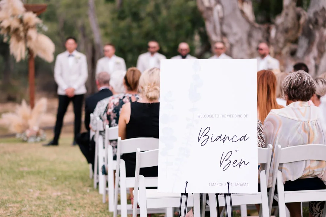 Outdoor wedding ceremony with guests seated in white chairs, large white wedding sign with the names Bianca and Ben, officiant standing in front of a natural backdrop of trees and rocks.