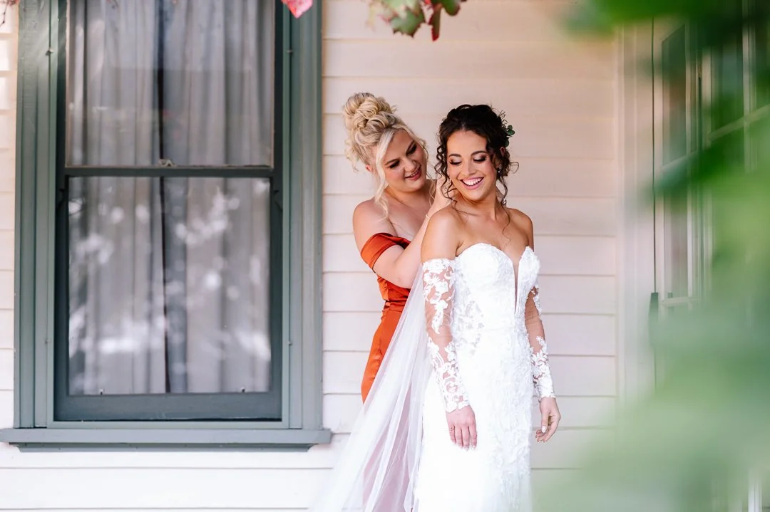 A bride in a white lace wedding gown standing on a porch, being helped with her dress by a woman in an orange dress.