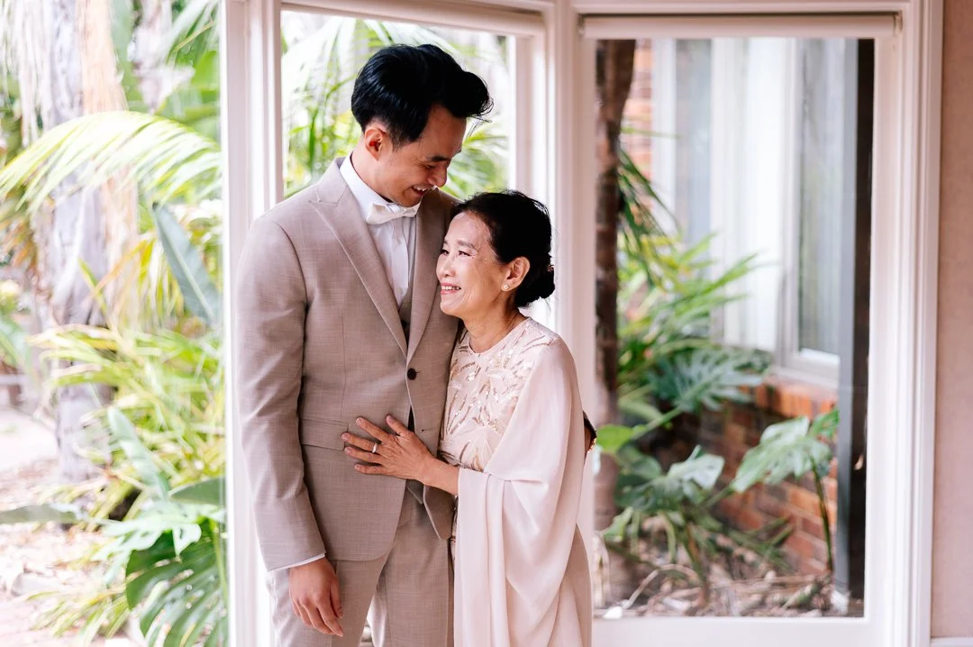 A young man and an elderly woman are smiling and embracing each other inside a room with large windows, with greenery visible outside.