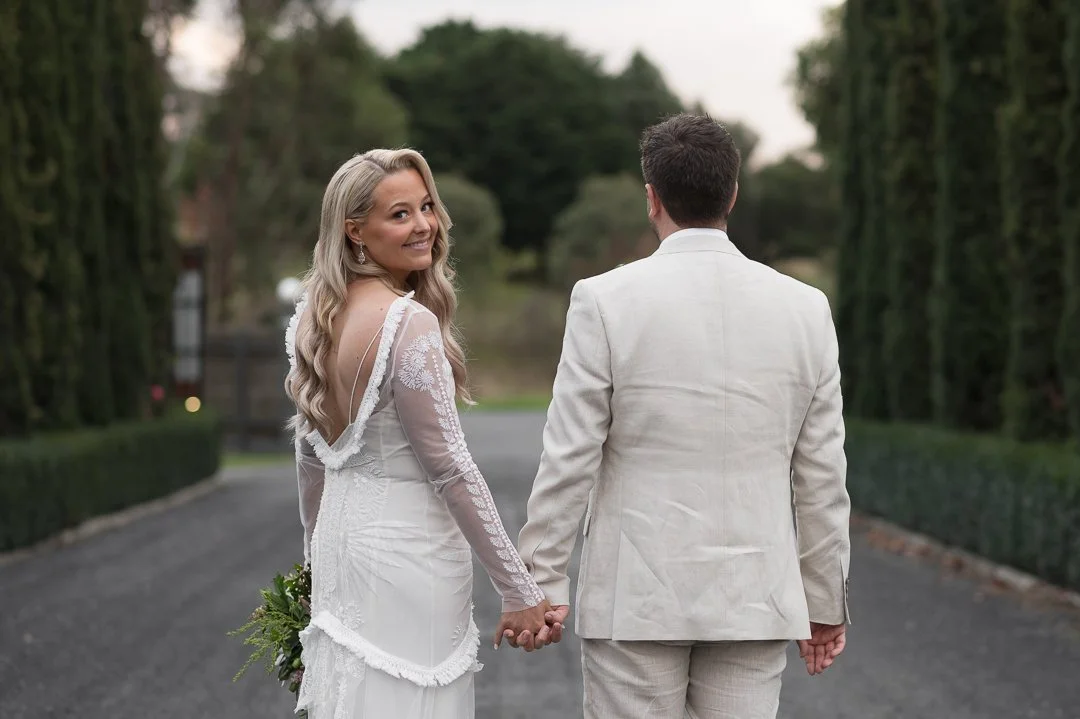 A couple dressed in wedding attire holding hands and walking outdoors on a tree-lined path