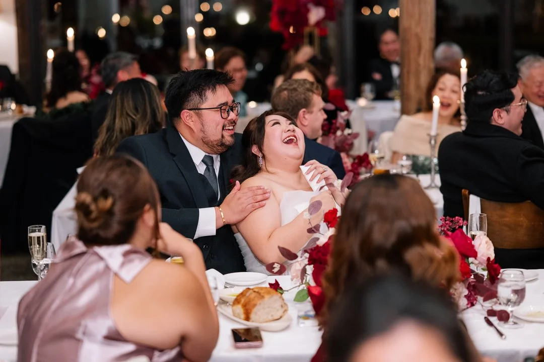 People sitting at a decorated wedding reception table, smiling and laughing, with candles and floral arrangements.