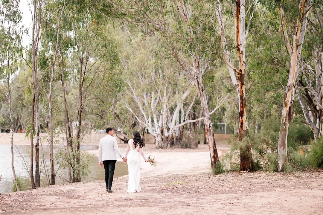 A bride and groom walking hand-in-hand in a wooded outdoor setting, with trees and a body of water nearby.