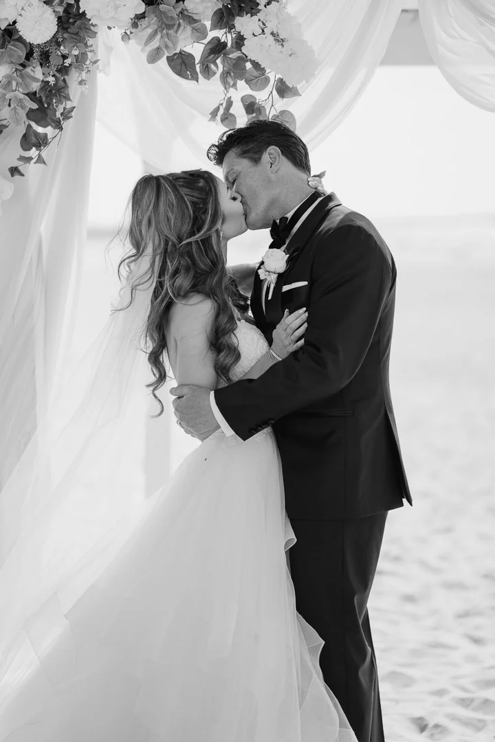 A black-and-white photo of a bride and groom sharing a kiss during their wedding ceremony outdoors near the water, under a decorated arch with flowers and greenery.