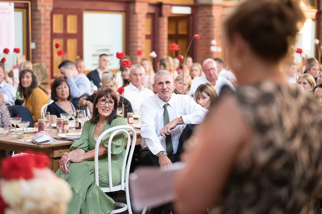 A woman giving a toast at a wedding reception, with guests seated at tables decorated with red flowers in a brick-walled venue.