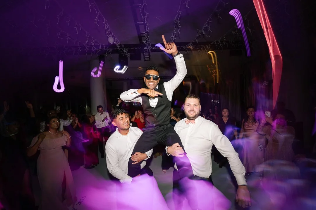 People dancing at a party, with three men in the foreground, one wearing sunglasses and lifting his hand in the air, surrounded by colorful lights and a lively crowd.