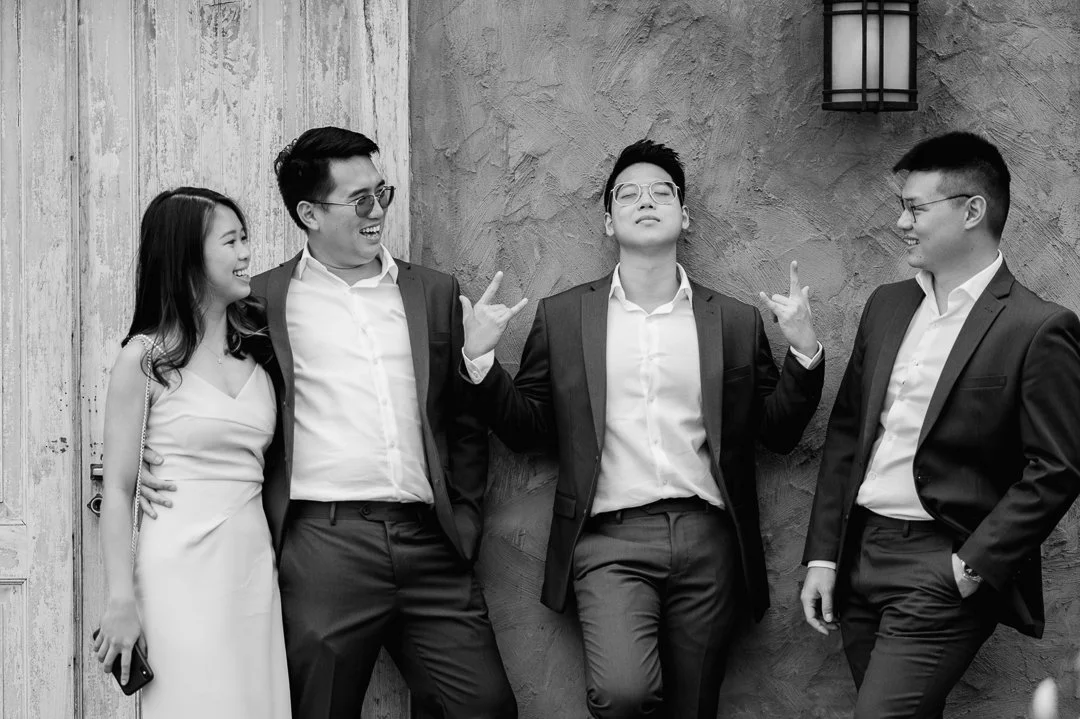 Five friends in formal attire standing against a textured wall, with one person making a peace sign and the others smiling.