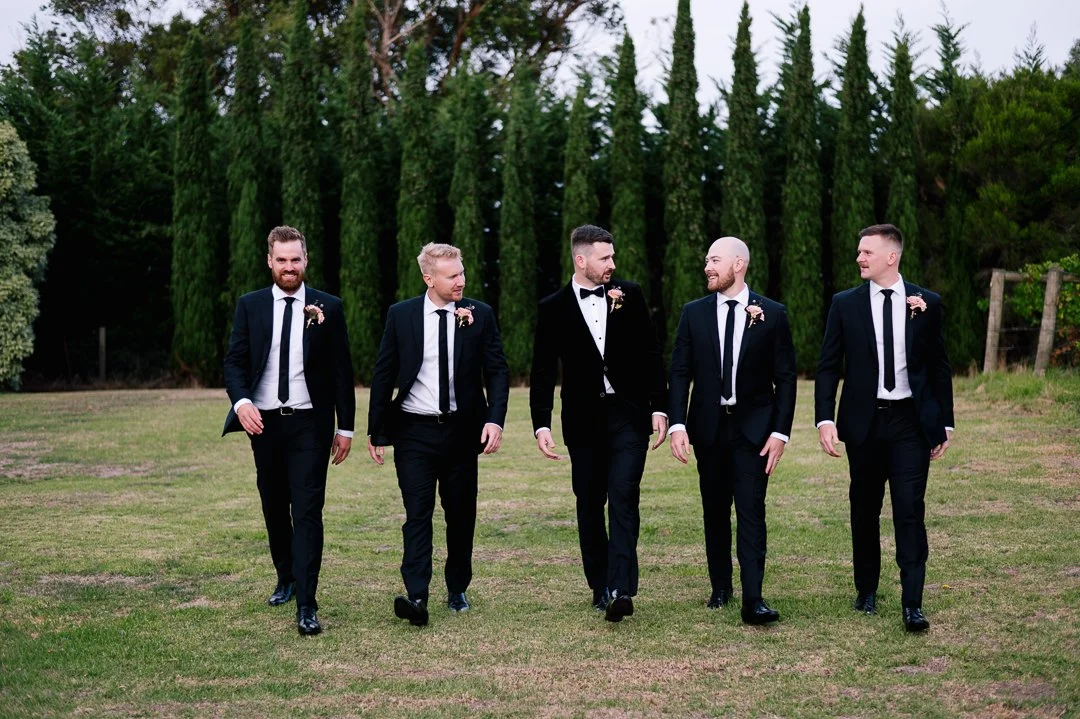 Group of five men in tuxedos walking outdoors in a green, wooded area, likely at a wedding or formal event.