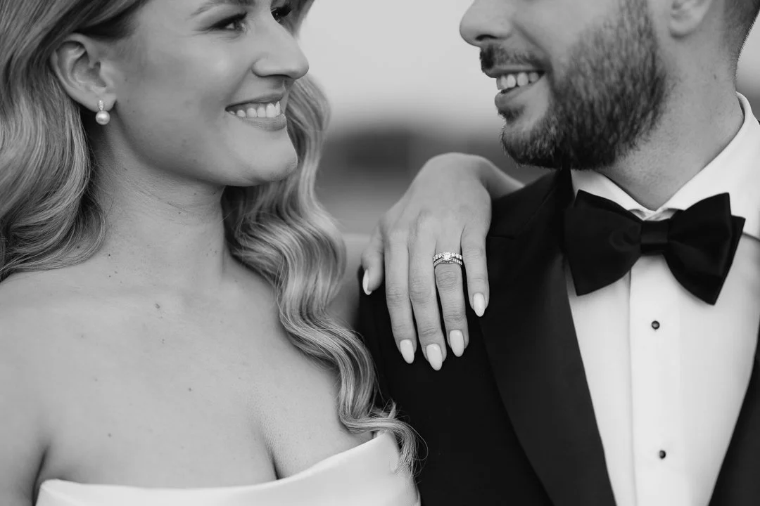 A woman and man dressed in formal wedding attire, smiling and looking at each other, with the woman resting her hand on the man's shoulder showing a wedding ring.