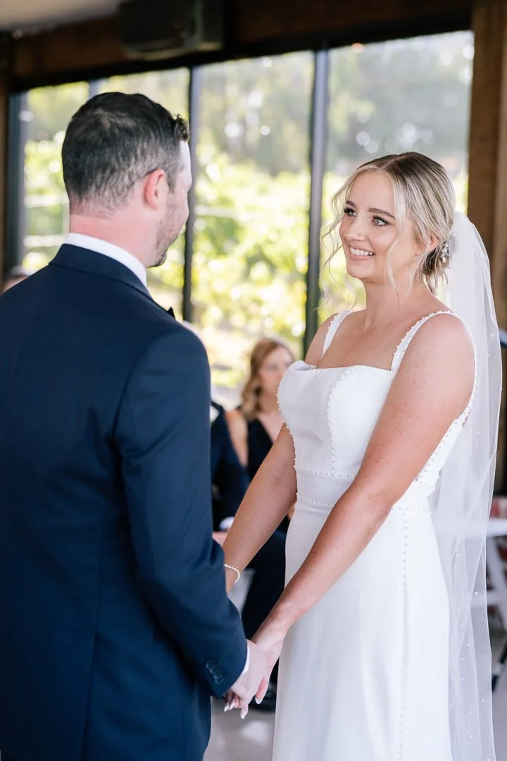 A bride and groom holding hands during their wedding ceremony, smiling at each other indoors with large windows showing greenery outside.