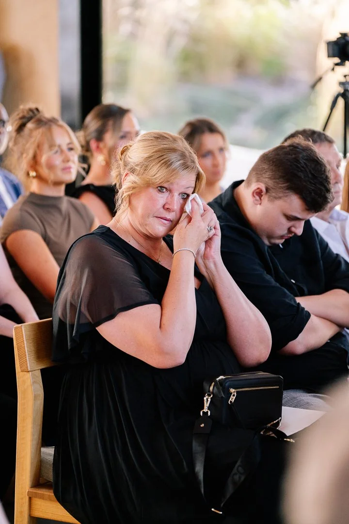 A woman with blonde hair sitting in a crowd, wiping tears from her eyes, with others seated around her and a camera on a tripod in the background.