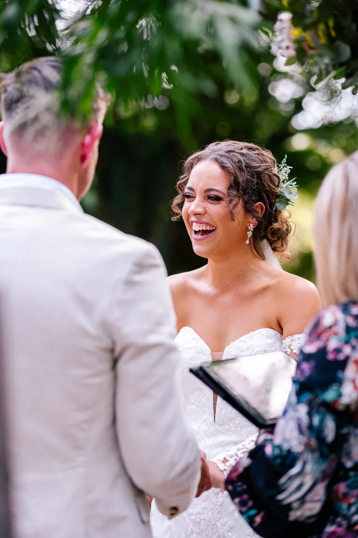 A bride with curly hair, wearing a strapless white wedding dress, smiling and laughing during her wedding ceremony outdoors, with a person in a white suit and an officiant holding a tablet.