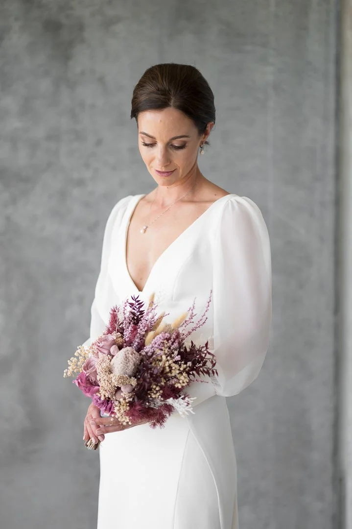 A woman in a white dress holding a pink and purple floral bouquet, standing against a gray background.