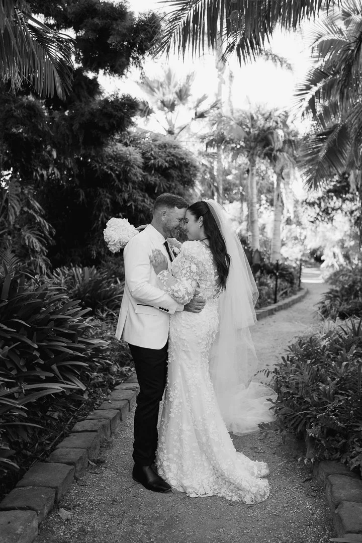 Black and white photo of a newlywed couple embracing outdoors on a garden path surrounded by tropical plants and palm trees.