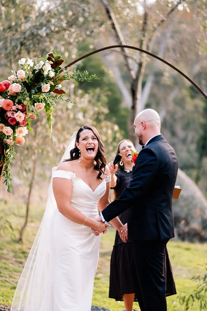 A bride and groom holding hands during their wedding ceremony outdoors, with an officiant behind them, under a floral arch, in a wooded area.