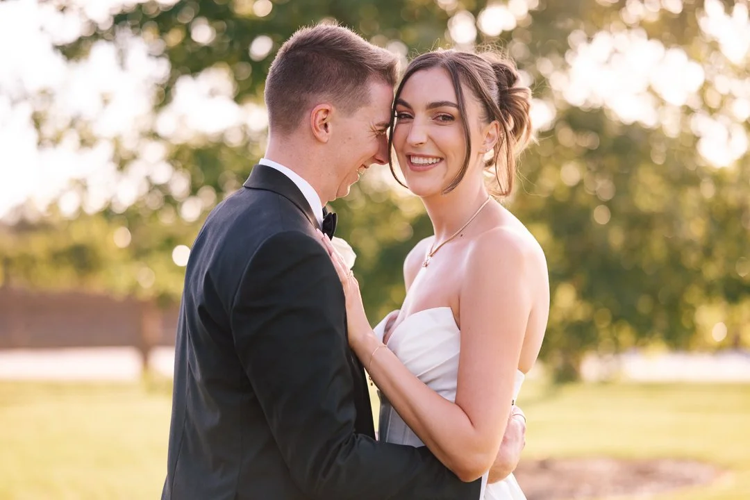 A newlywed couple smiling and embracing outdoors in a park at sunset, with trees and warm light in the background.