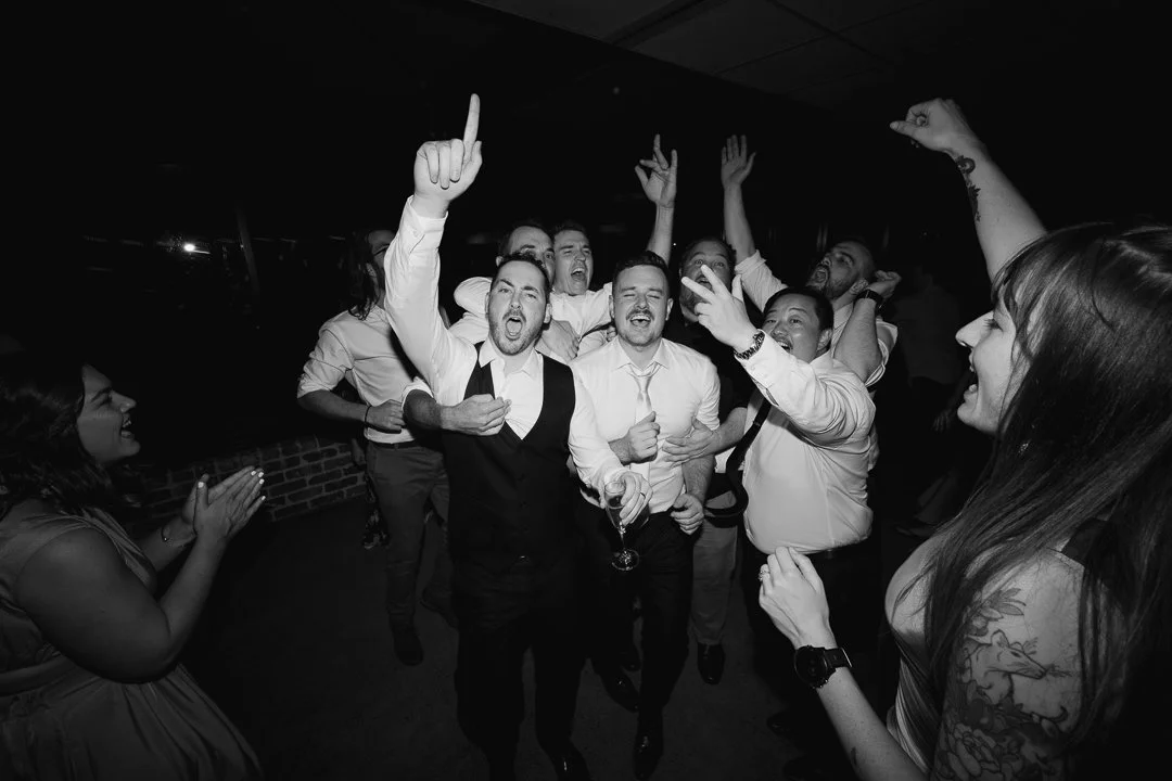 Group of people celebrating and dancing at a party or event, some with drinks in hand, smiling and raising their hands.